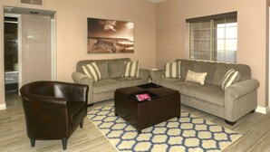 A living room with a brown leather chair, two grey couches with striped pillows, a brown ottoman with a book, and a patterned rug. - Beautiful home with 2 masters! (Lake Havasu City)
