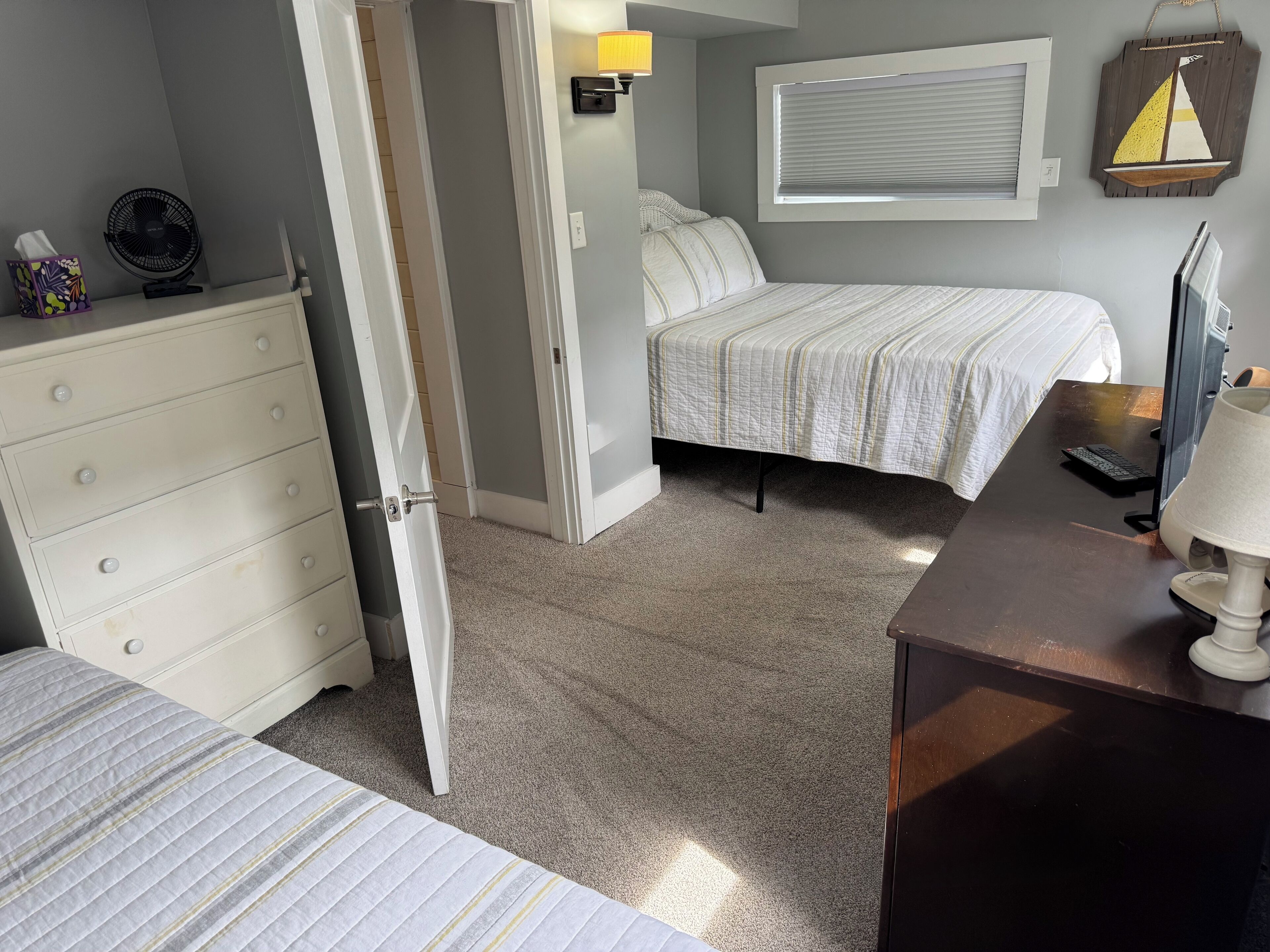 A bedroom with a bed, a dresser, a desk, a lamp, a wall-mounted fan, a window with blinds, and a decorative sailboat.