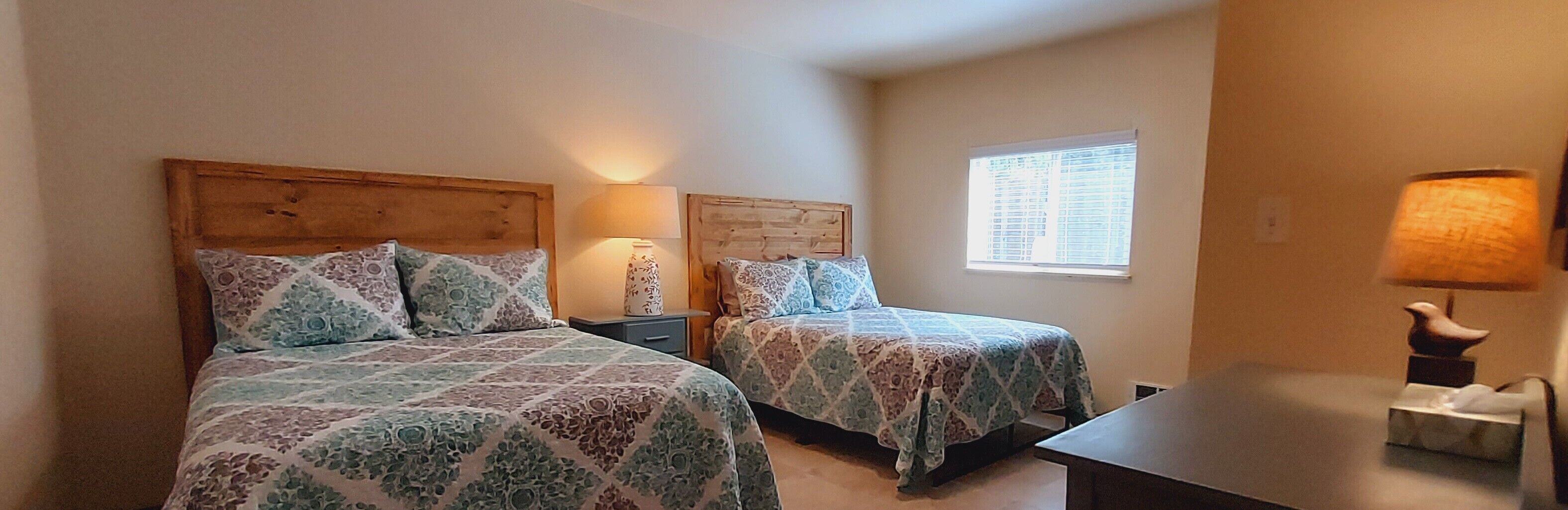 Un dormitorio con dos camas, cada una con cabecero de madera y ropa de cama estampada. Hay una mesita de noche con una lámpara entre las camas y una ventana con persianas en la pared derecha.