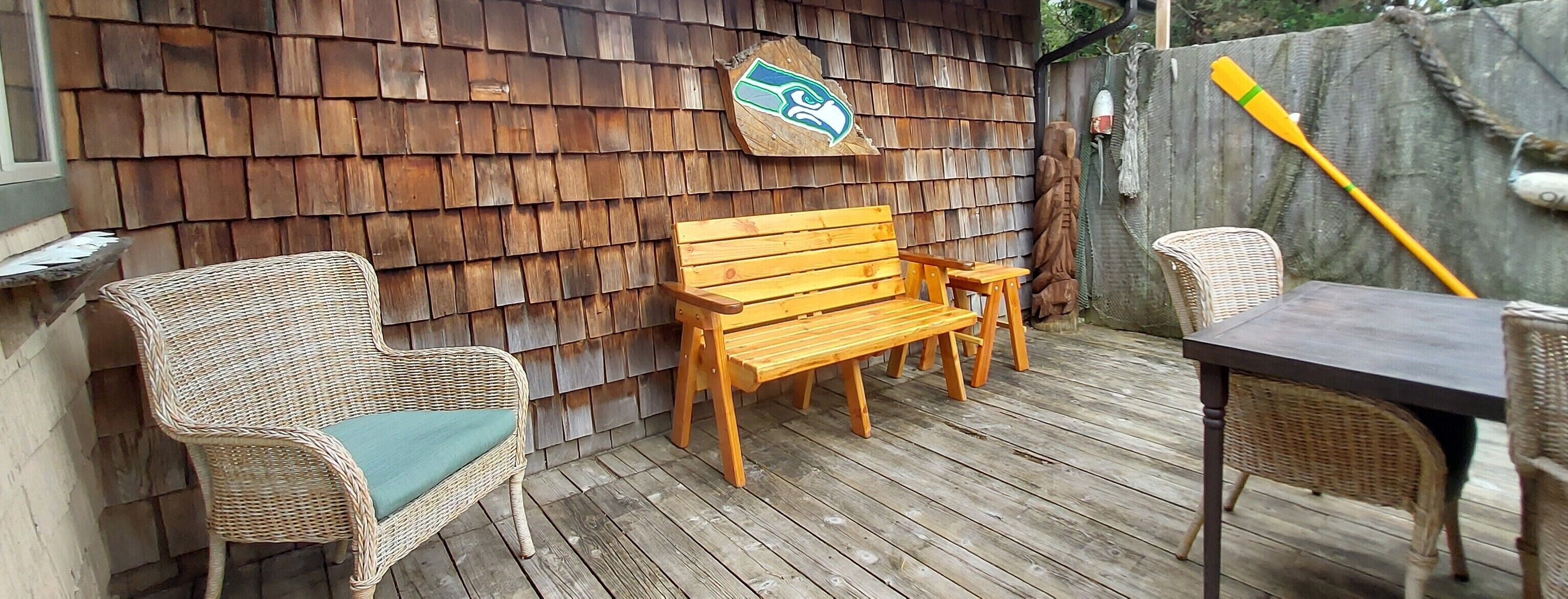 A wooden deck with a bench and wicker chairs, a table, and a wooden wall with a logo.