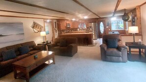A cozy living room with a sofa, armchairs, and a coffee table. A kitchen area with a refrigerator and sink is visible in the background.