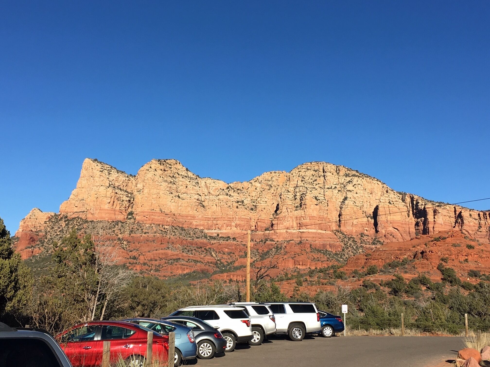 A parking area with several cars, a clear blue sky, and a large red rock formation in the background.