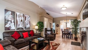 A modern living room with a brown sofa, red cushions, and abstract wall art. There's a dining area with a table and chairs, a kitchen in the background, and a large potted plant. - Little taste of Heaven, 5 min walk to pool/hot tub, close to hikes & restaurants (Sedona)