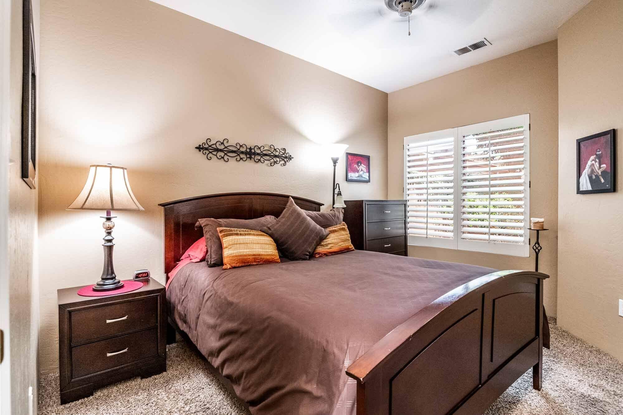 Un dormitorio con cama, mesitas de noche, lámpara, cómoda, ventana con persianas y decoración en la pared.