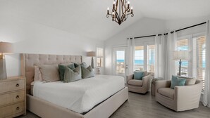 Une chambre spacieuse avec un grand lit, deux fauteuils, un lustre et une vue sur la mer grâce à de grandes fenêtres.