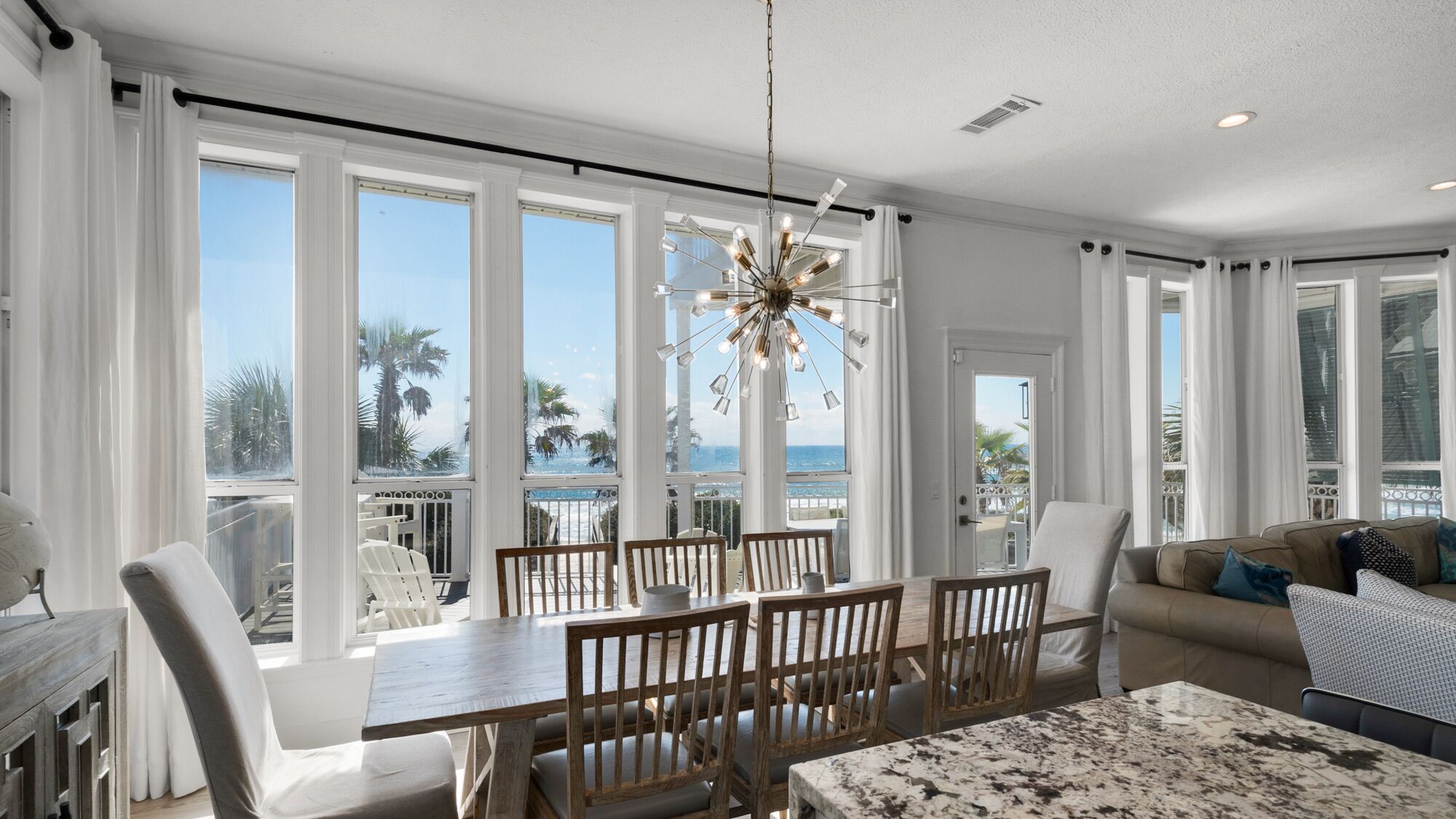 Ein geräumiges Wohnzimmer mit großem Esstisch und Stühlen, einer modernen Kronleuchter und großen Fenstern mit Blick auf das Meer und Palmen.