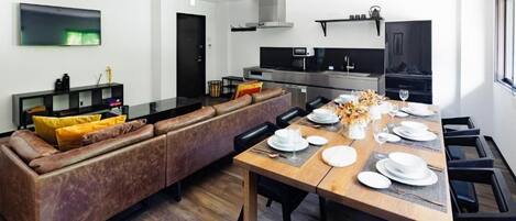 A modern kitchen with a built-in oven and microwave, a dining area with a wooden table set for four, and a brown leather sofa with yellow cushions.