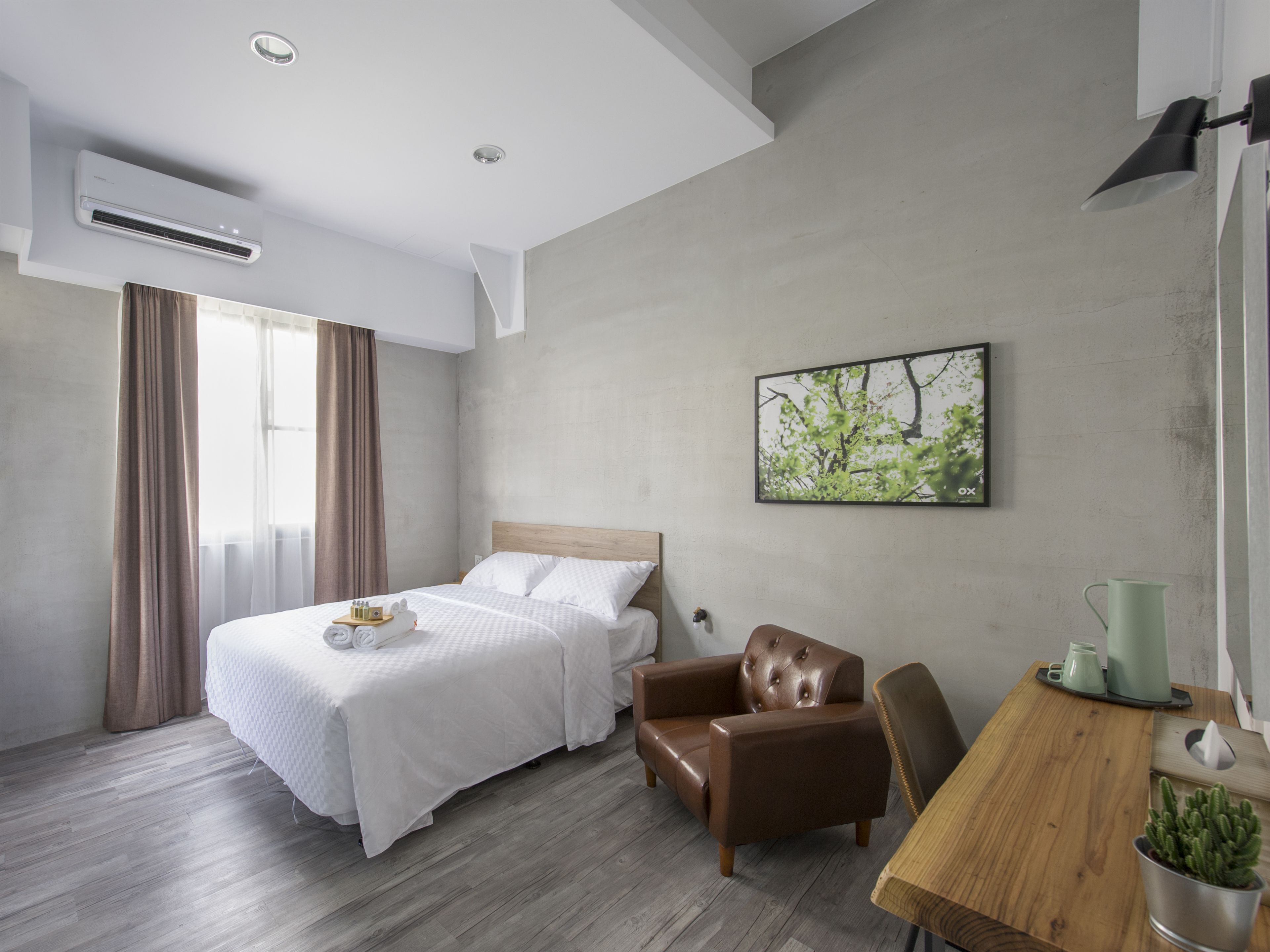 A modern hotel room with a bed, a leather armchair, a wooden table, and a wall-mounted picture.
