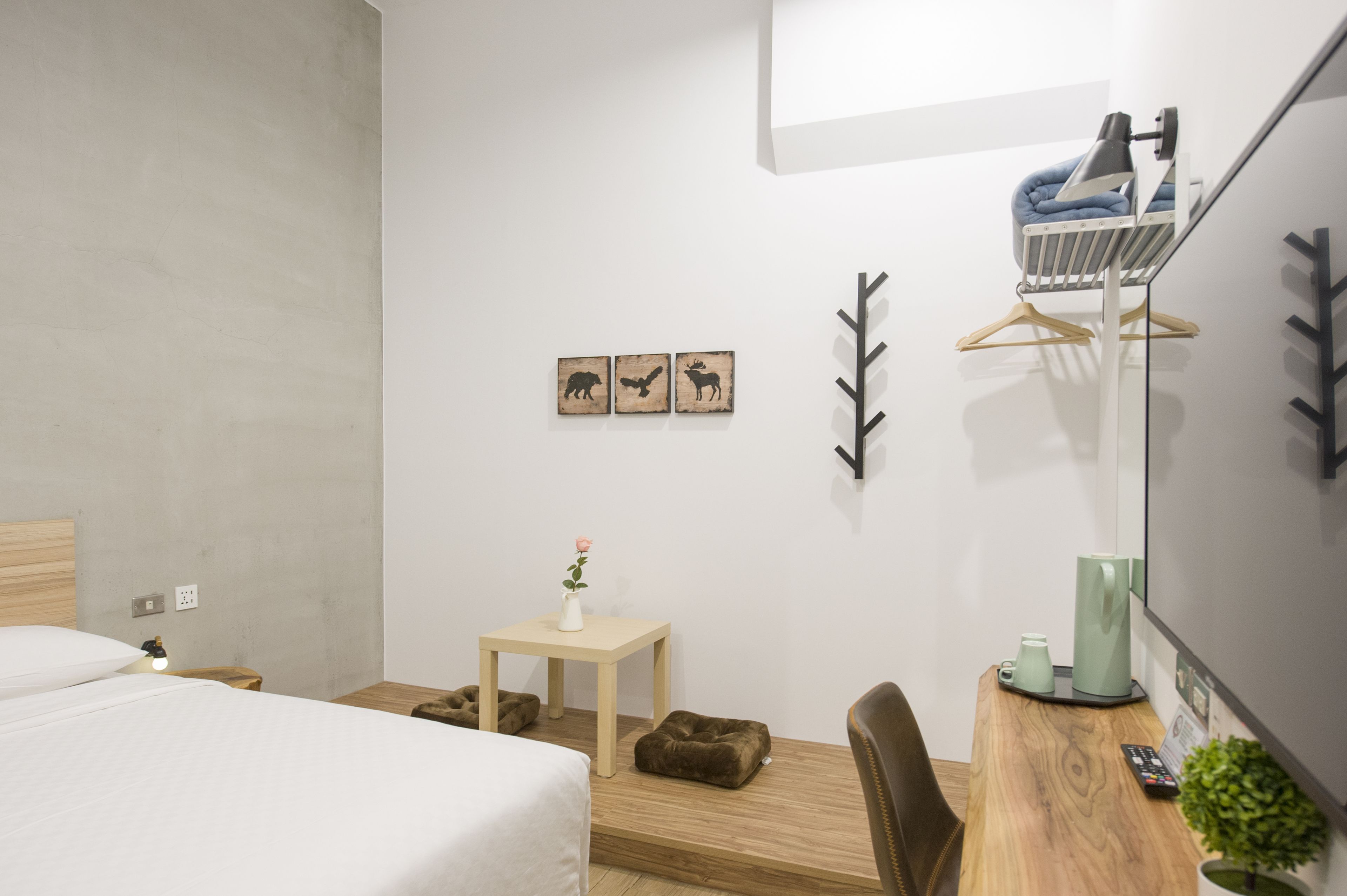 A hotel room with a bed, bedside table with a vase of flowers, a desk with a coffee maker, a chair, and wall decorations including animal silhouettes and a leaf pattern.