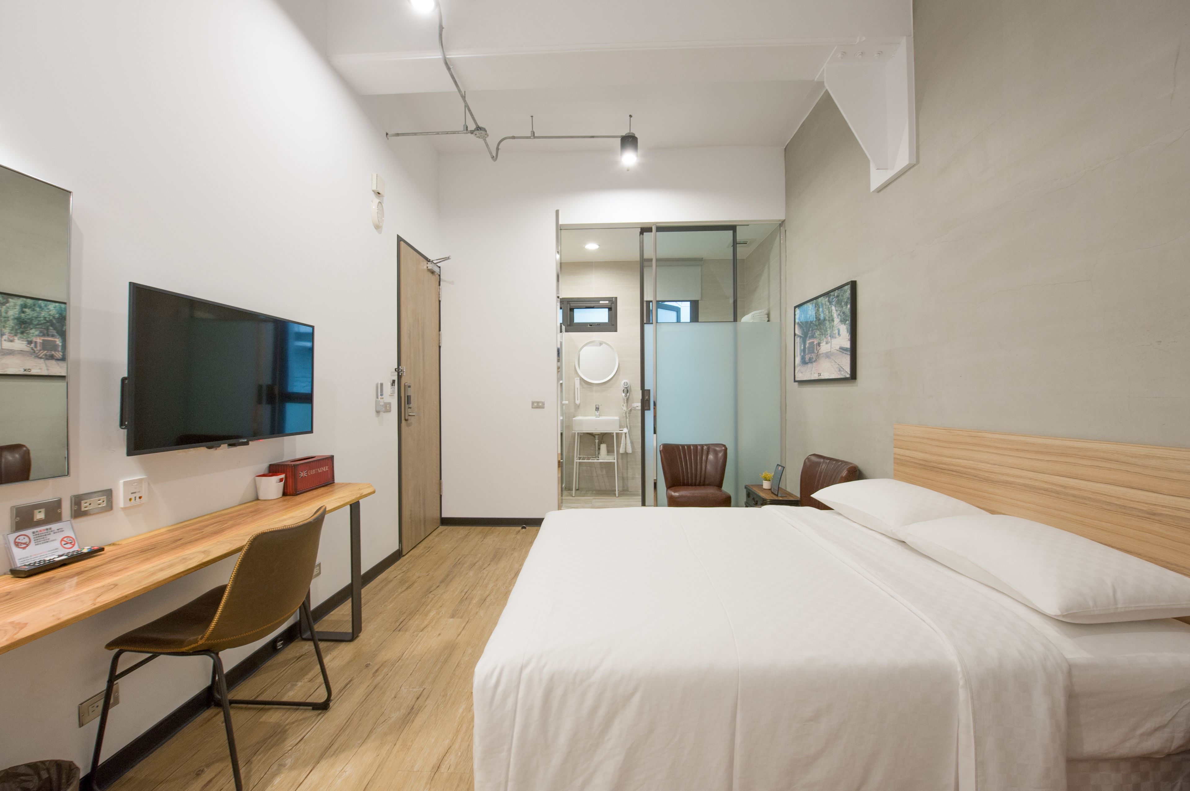 A modern hotel room with a wooden desk, a bed with white linens, a television, and a bathroom visible through a glass door.