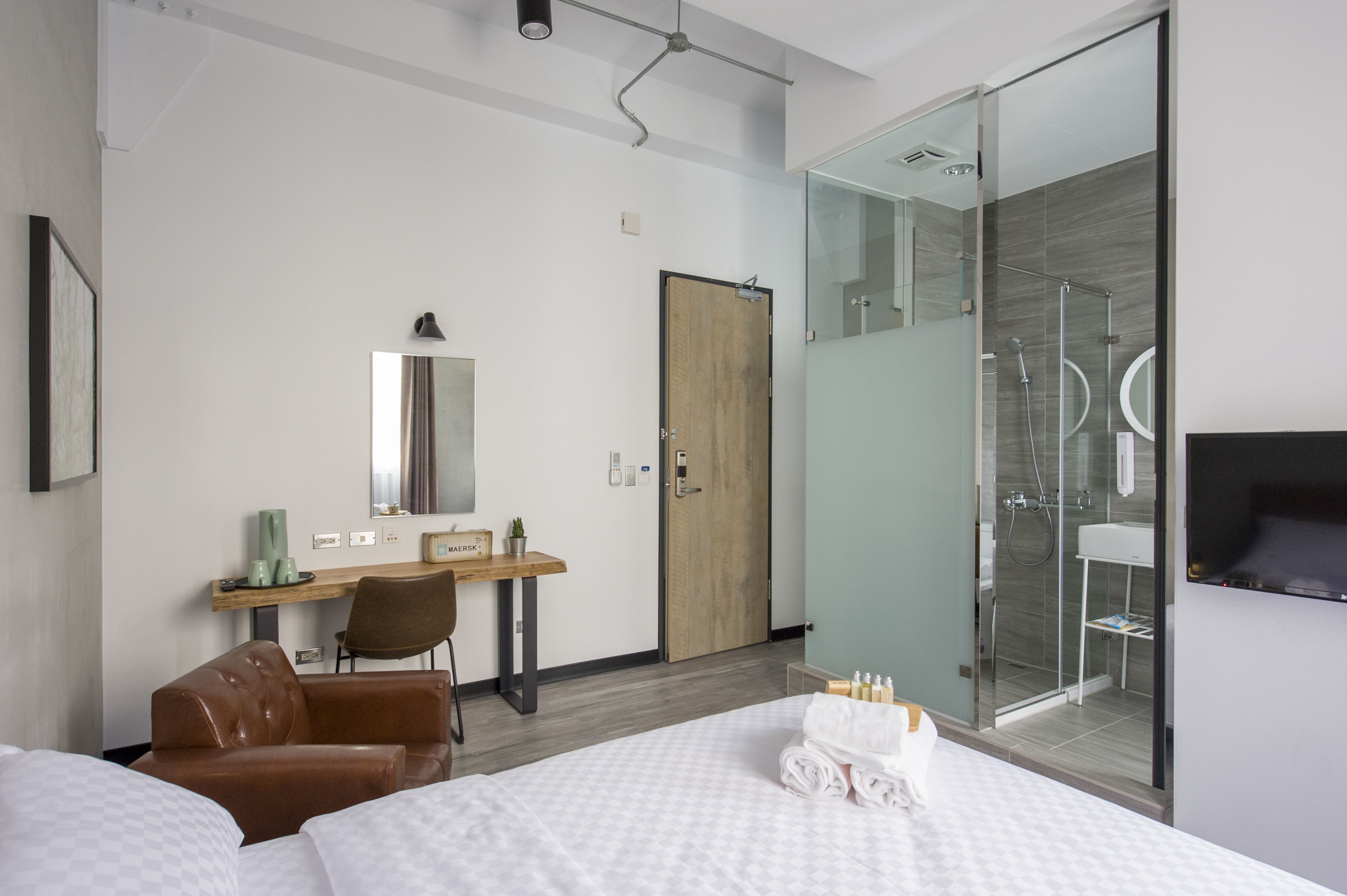 A modern hotel room with a bed, a desk, a chair, a mirror, a door, and a shower area.