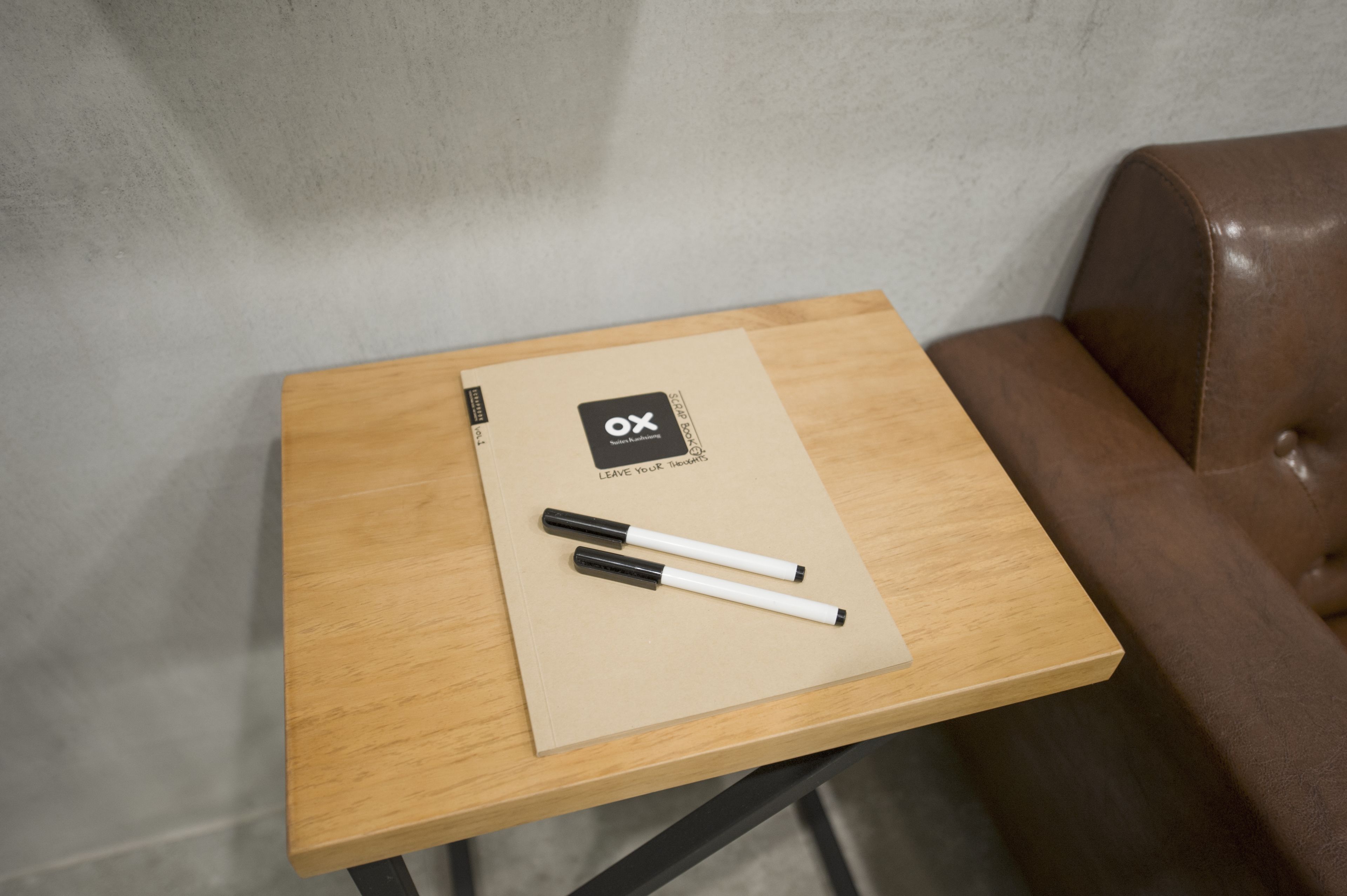 Une petite table en bois, sur laquelle reposent un classeur beige et deux stylos, est placée contre un mur gris, à côté d’un canapé en cuir marron.