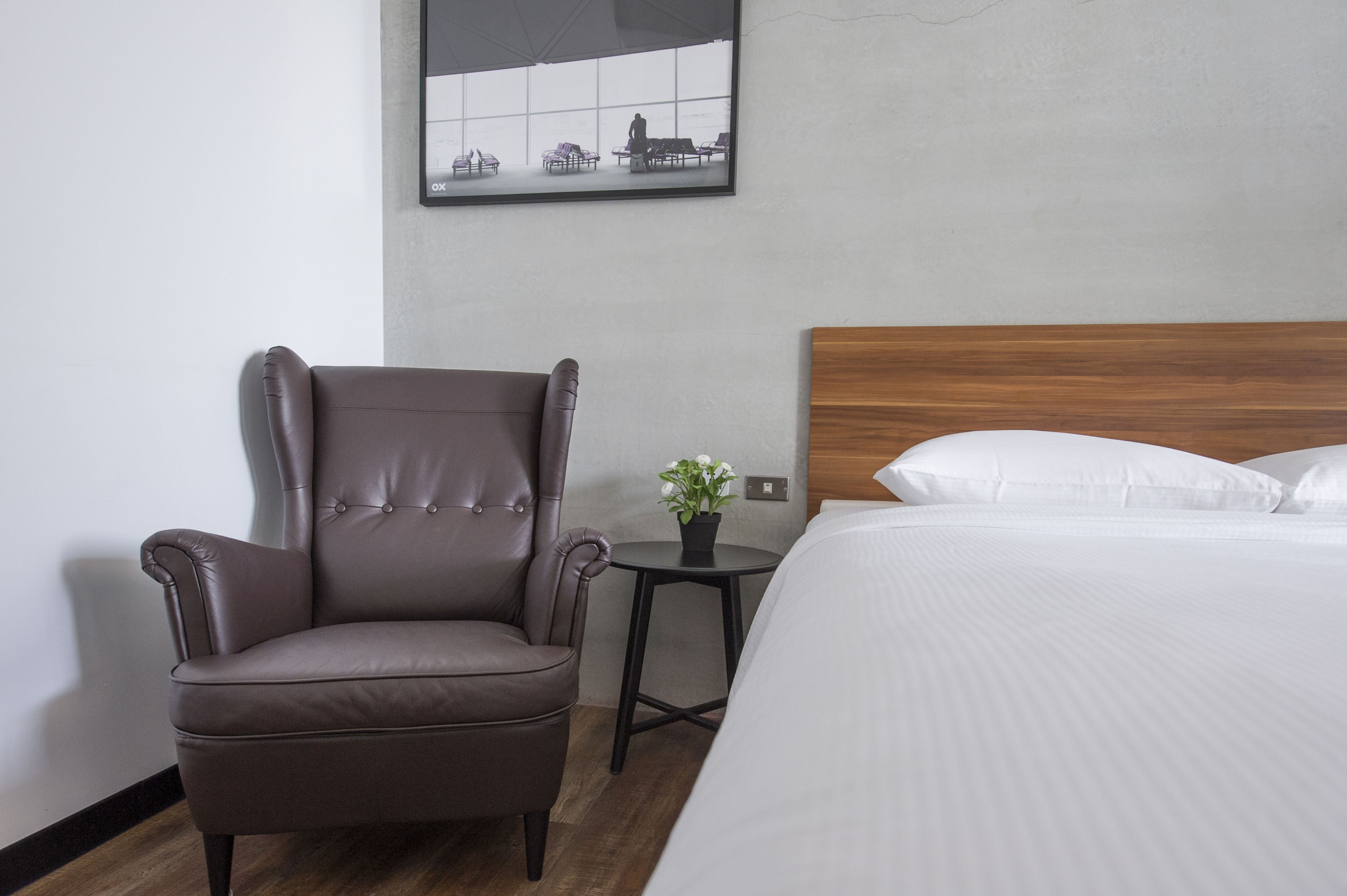 A hotel room with a large bed, a brown leather armchair, a small table with a plant, and a framed picture on the wall.