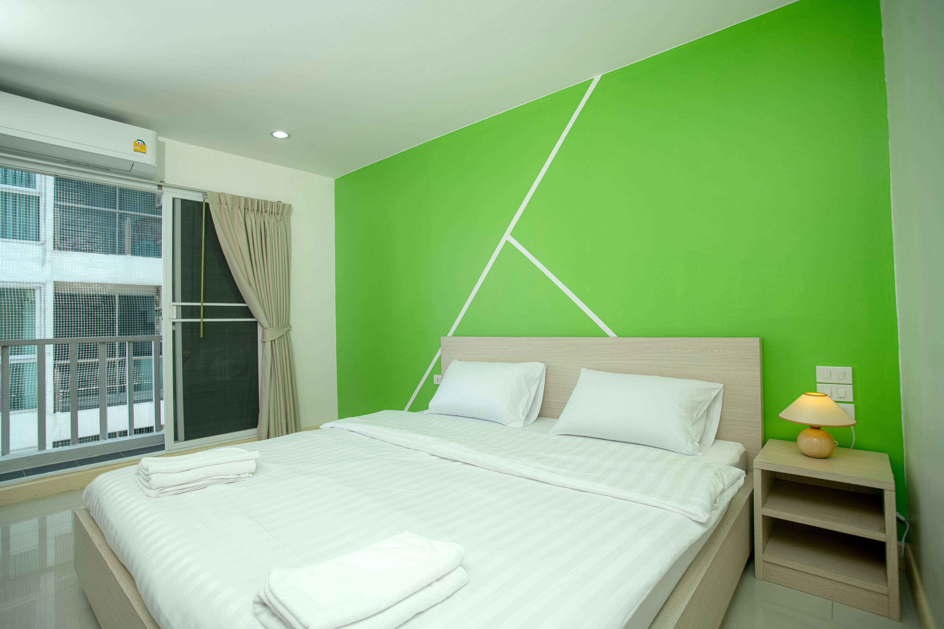 A modern hotel room with a large bed, a green accent wall, a window with a view, and a bedside table with a lamp.