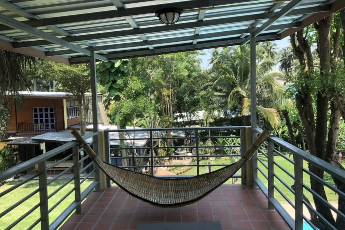 Área externa coberta com uma rede, guarda-corpo de metal e vista para um jardim com árvores e um edifício ao fundo.