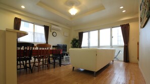 A spacious living room with a sofa, dining table, and chairs. There is a large window with curtains, an air conditioning unit, and a clock on the wall. - JQ Villa Okinawa Yaka (Kin)