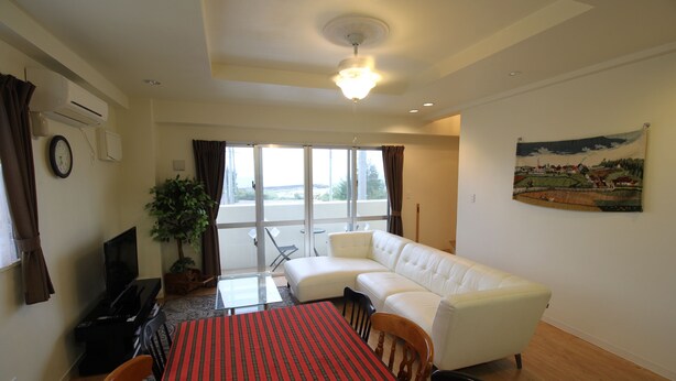 A living room with a white sofa, a dining table with a red tablecloth, a potted plant, and a large window with curtains. - JQ Villa Okinawa Yaka (Kin)