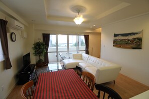 A living room with a white sofa, a dining table with a red tablecloth, a potted plant, and a large window with curtains.