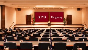 Ein Konferenzraum mit Reihen von Tischen und Stühlen, eine Bühne mit Rednerpult und ein großer Bildschirm, der "SPS Kingsway" zeigt.