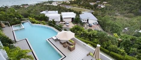 A rooftop pool with lounge chairs and an umbrella, surrounded by greenery and overlooking a coastal area with boats.