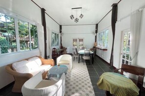 A sunlit room with a sofa, chairs, a dining table, and a view of greenery outside. - Khoom Kam Kaew (Phrao)
