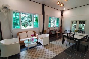 Ein Zimmer mit einer Couch, zwei Sesseln, einem kleinen Tisch, einem Esstisch mit Stühlen und großen Fenstern mit Blick ins Grüne.