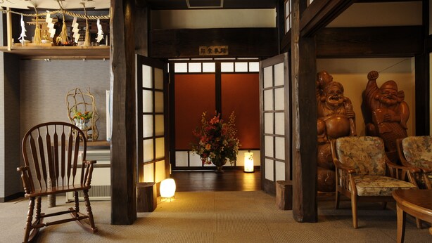 A traditional Japanese interior with tatami flooring, a wooden rocking chair, a flower arrangement, and a reception area with a sign. - Gensen no Yado Maruishi Ryokan (Hakuba)