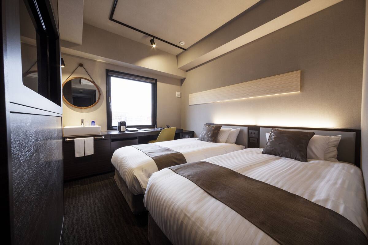 Une chambre d’hôtel moderne avec deux lits, un bureau, un grand miroir rond et une grande fenêtre.
