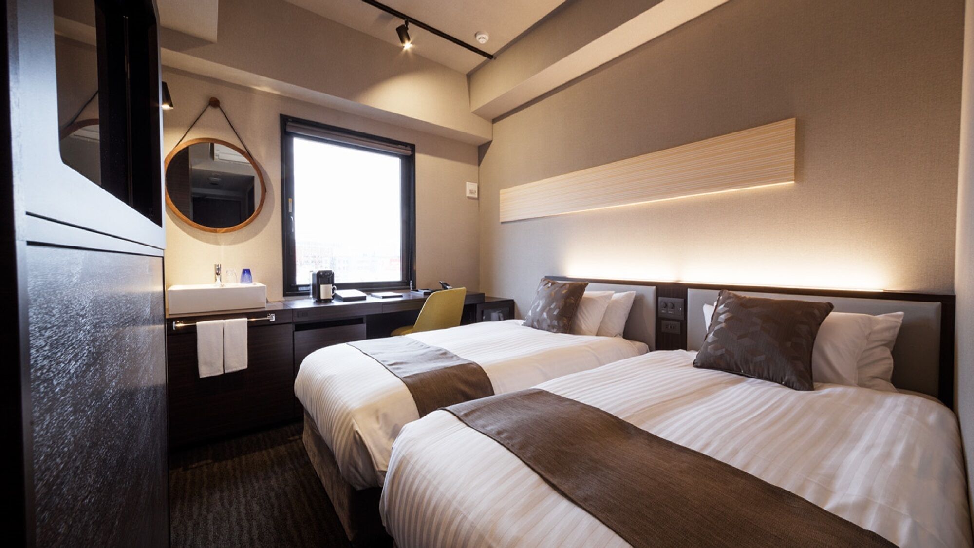 A hotel room with two beds, a desk, a mirror, a window, and a wall-mounted light fixture.