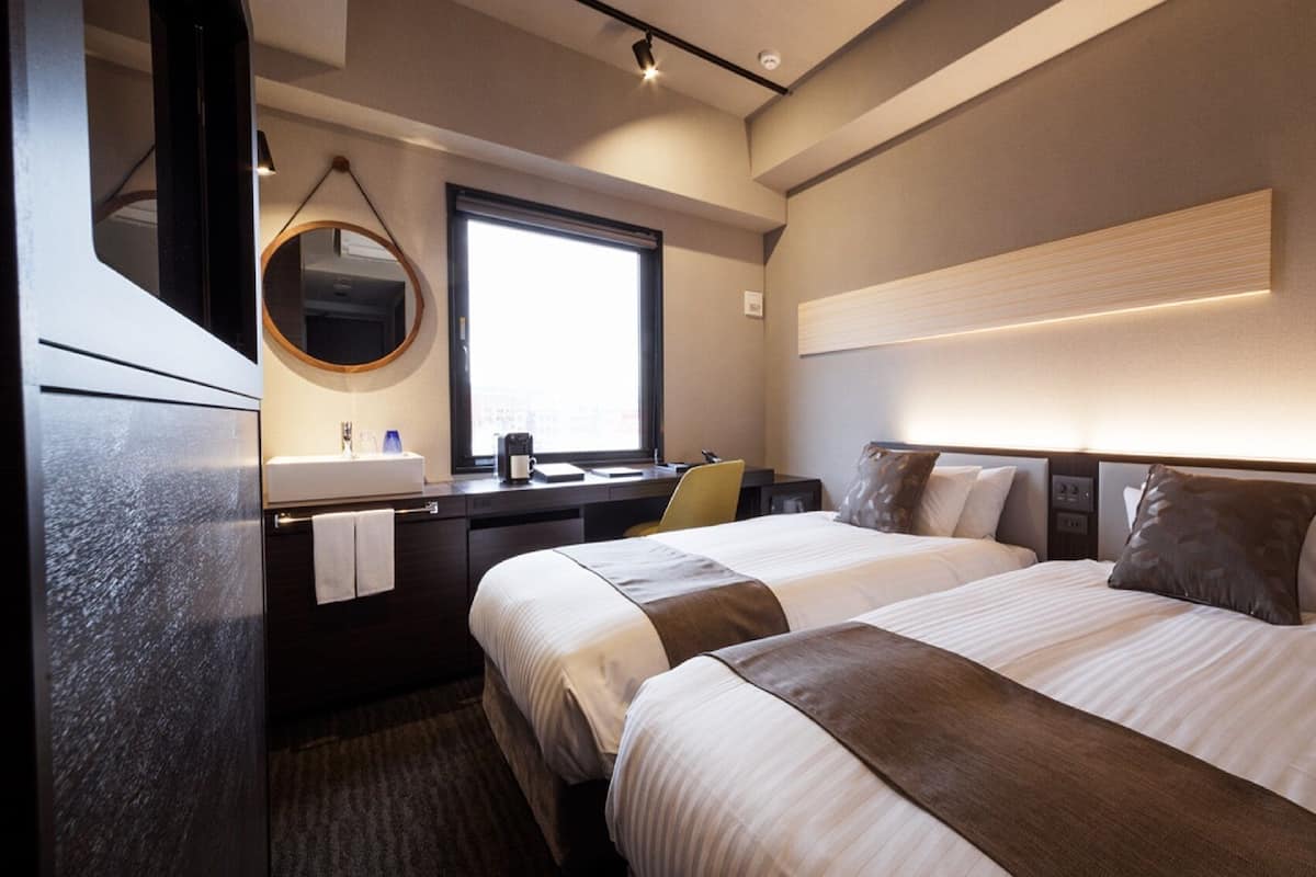 Une chambre d’hôtel avec deux lits, un bureau, un miroir, une fenêtre et un luminaire fixé au mur.