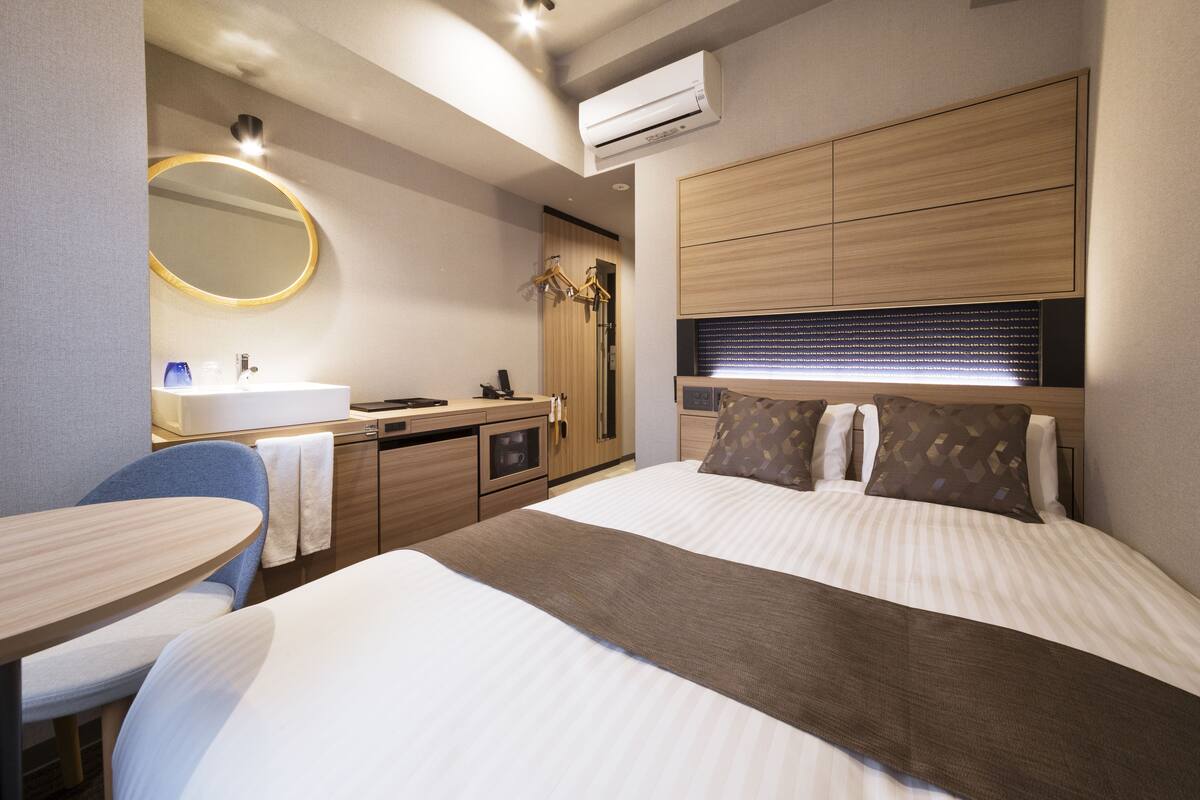 Une chambre d’hôtel moderne dotée d’un grand lit, d’une table de chevet, d’un miroir rond, d’une armoire intégrée et d’un petit bureau avec une chaise.