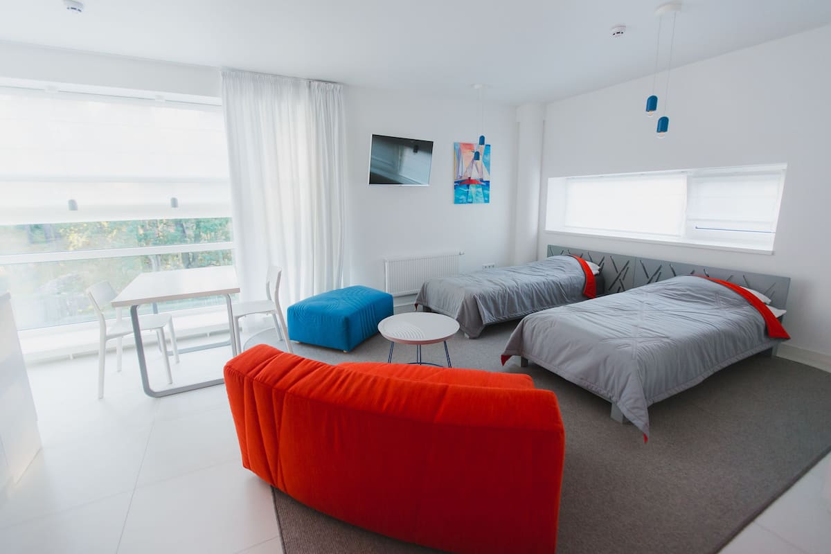 A modern hotel room with a large window, a dining table, two beds, and a bright orange sofa.