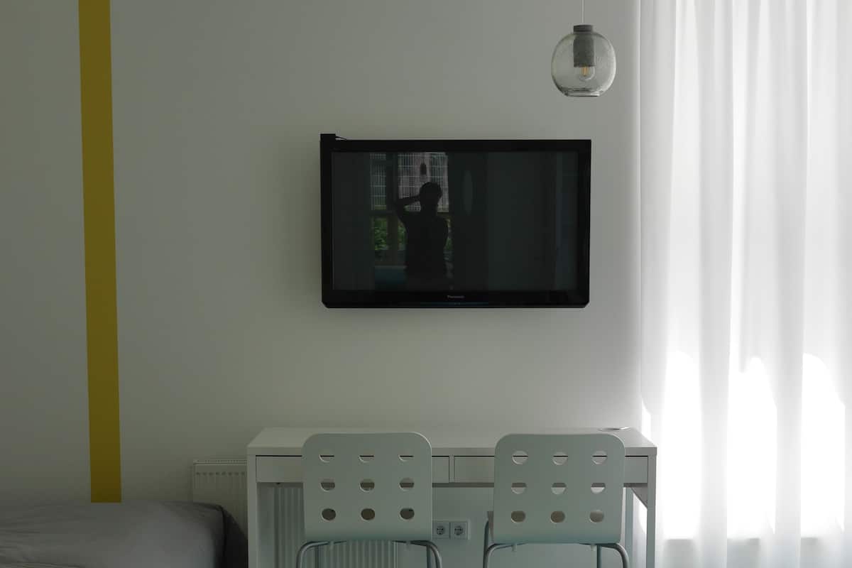 A room with a bed, a desk with two chairs, a television, and a window with curtains.