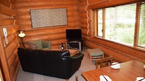 Interior de una cabaña de troncos con un sofá de cuero negro, una mesa de comedor de madera para cuatro personas, un televisor y una lámpara.