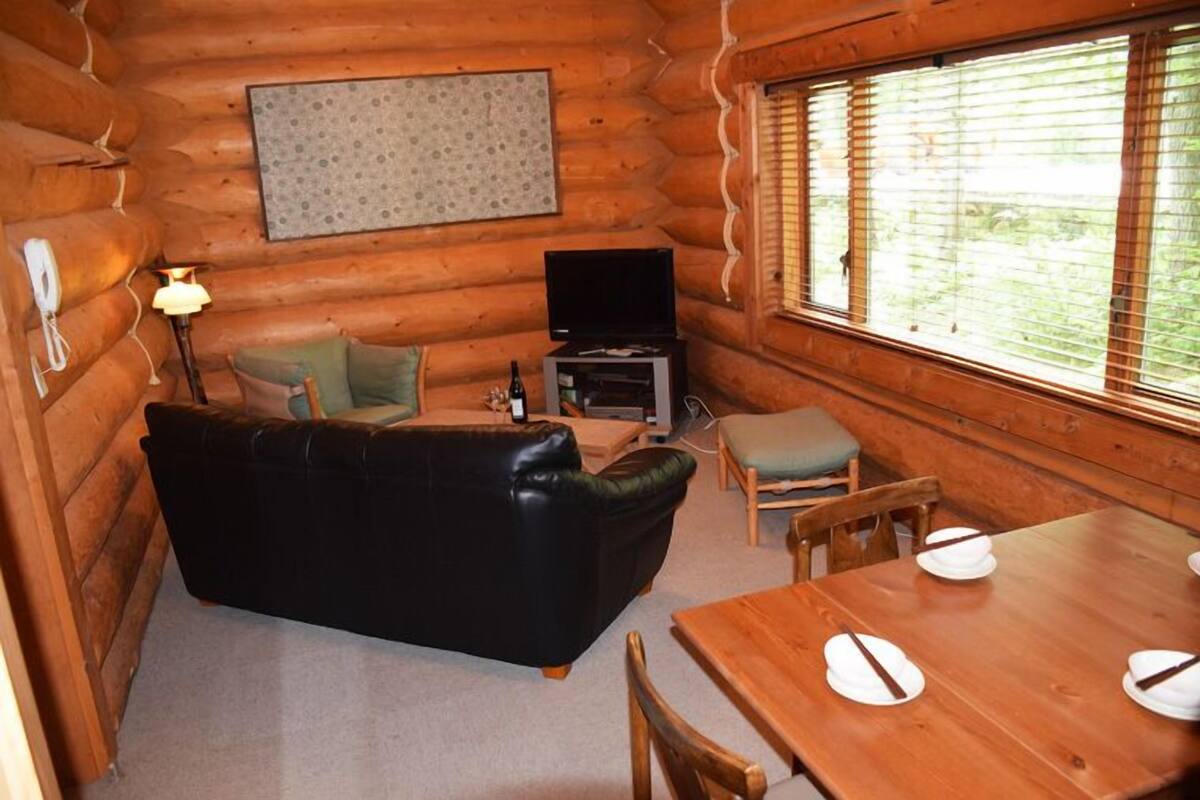 Interior de una cabaña de troncos con un sofá de cuero negro, una mesa de comedor de madera para cuatro personas, un televisor y una lámpara.