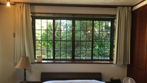 Un dormitorio con un ventanal grande, una cama, una mesita de noche, una lámpara y un ventilador de pie.