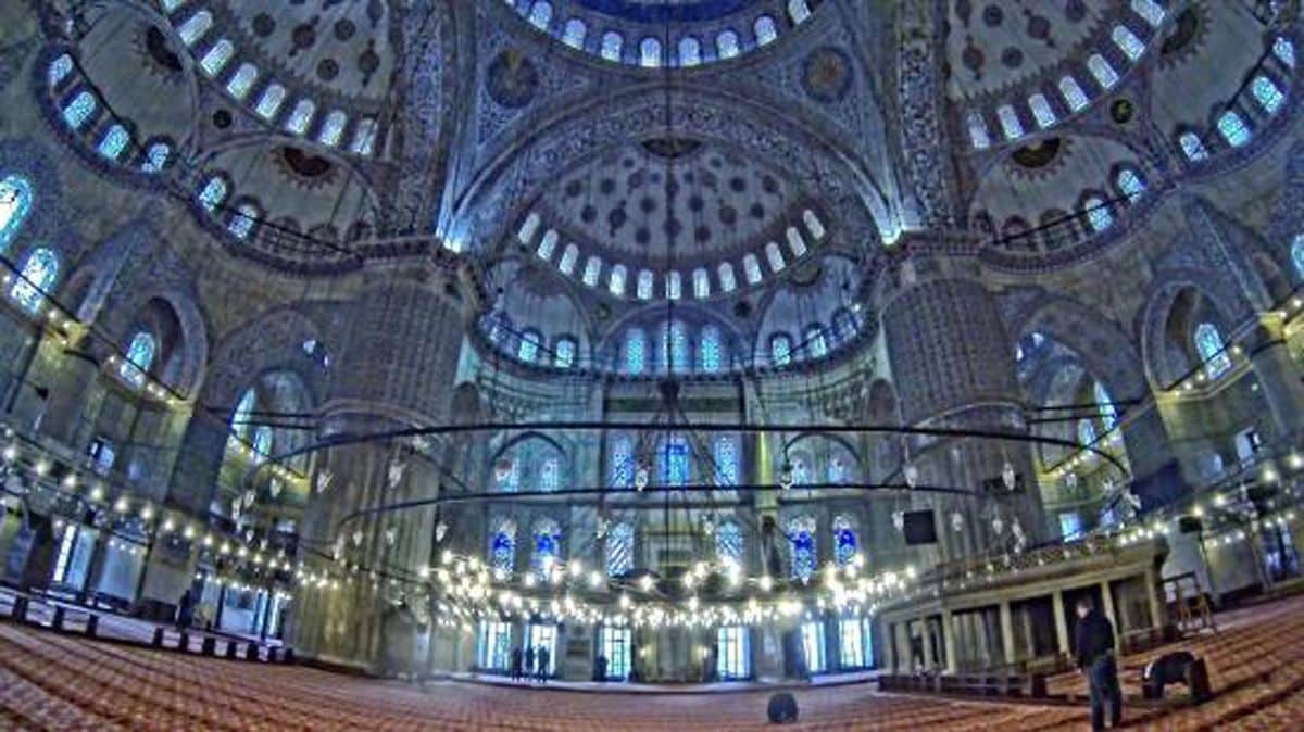 Vue intérieure d’un grand hall somptueux, doté d’un plafond élevé et présentant des détails architecturaux complexes ainsi que plusieurs niveaux d’arcades.