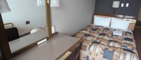 Habitación de hotel con cama, escritorio, silla, espejo y dos lámparas fijadas a la pared.