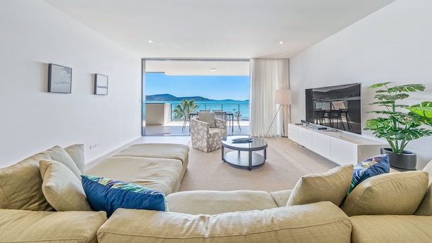 Een moderne woonkamer met een bank, een ronde salontafel en een televisie. Er is een balkon met uitzicht op zee en bergen.