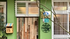 Entrada de un edificio con un muro verde, una puerta de madera y un ventanal con persiana blanca.