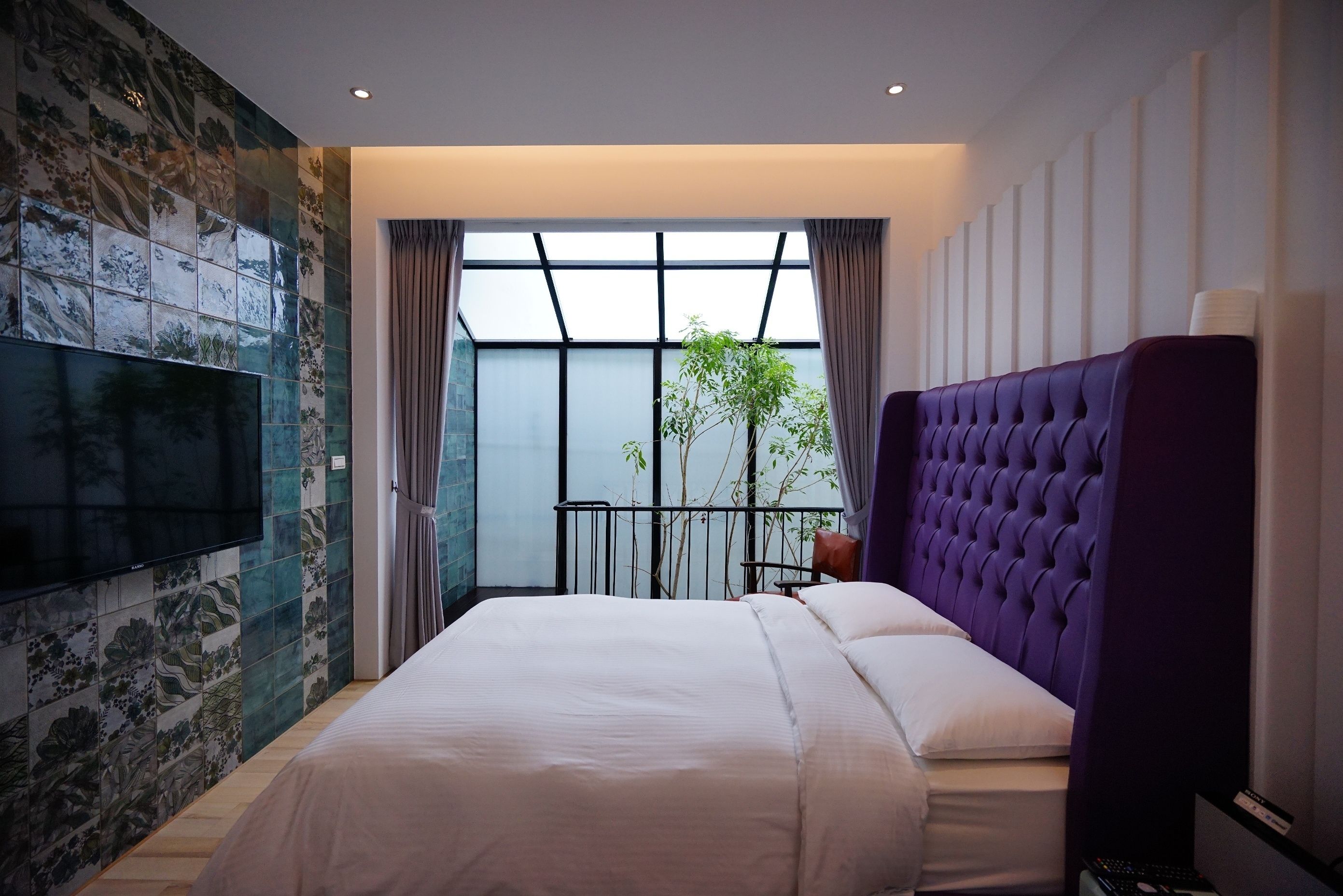 A modern hotel room with a large bed, a flat-screen TV mounted on the wall, a balcony with a view, and a purple headboard.
