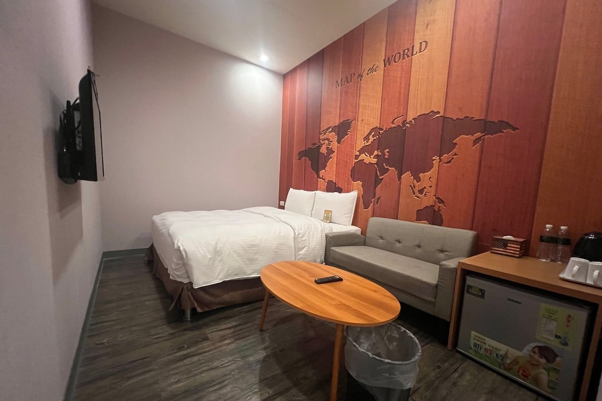 Una habitación de hotel con cama, sofá gris, pequeña mesa de madera, cubo de basura y televisor montado en la pared.