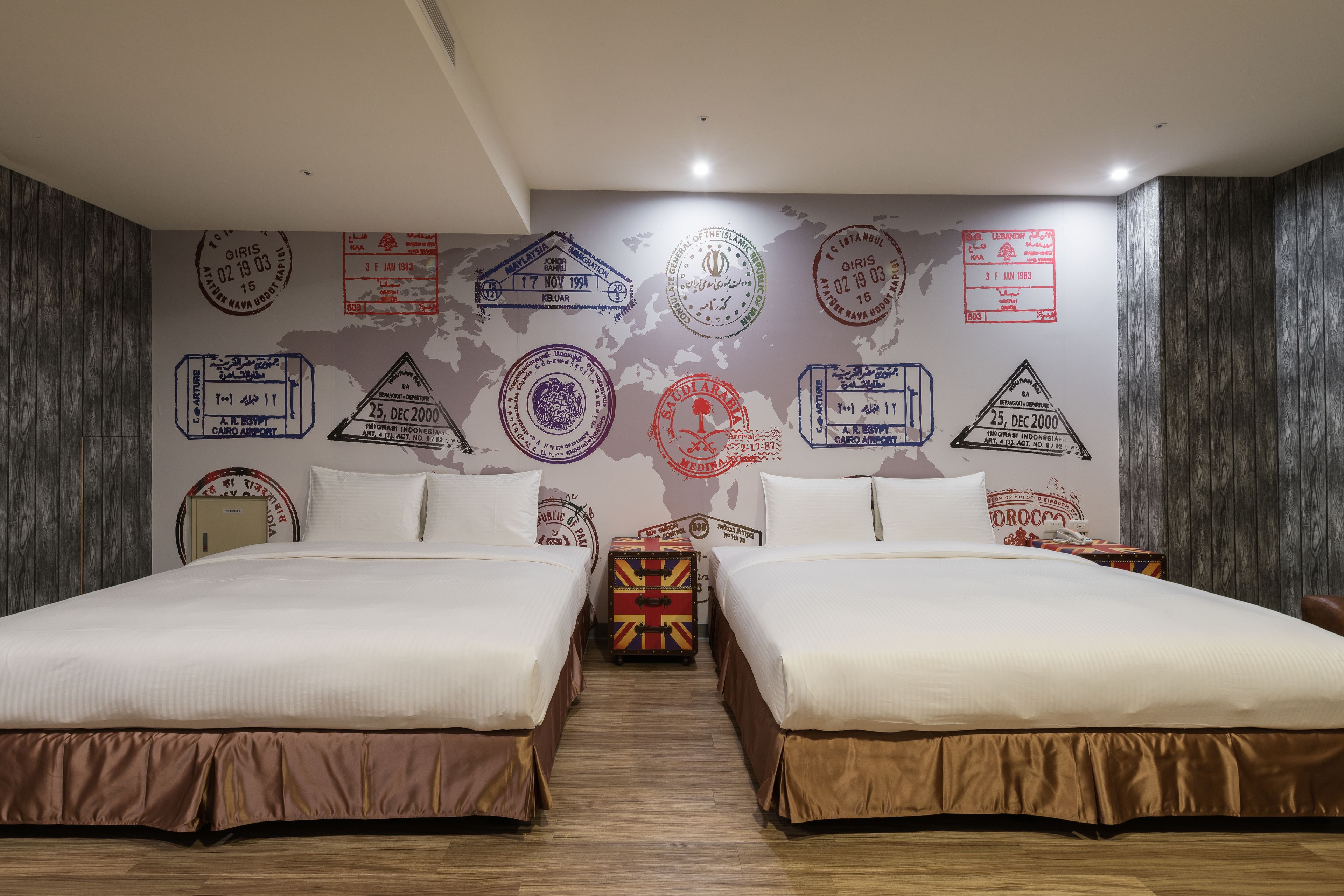 A hotel room with two beds, a wooden floor, and a wall decorated with various stamps and maps.