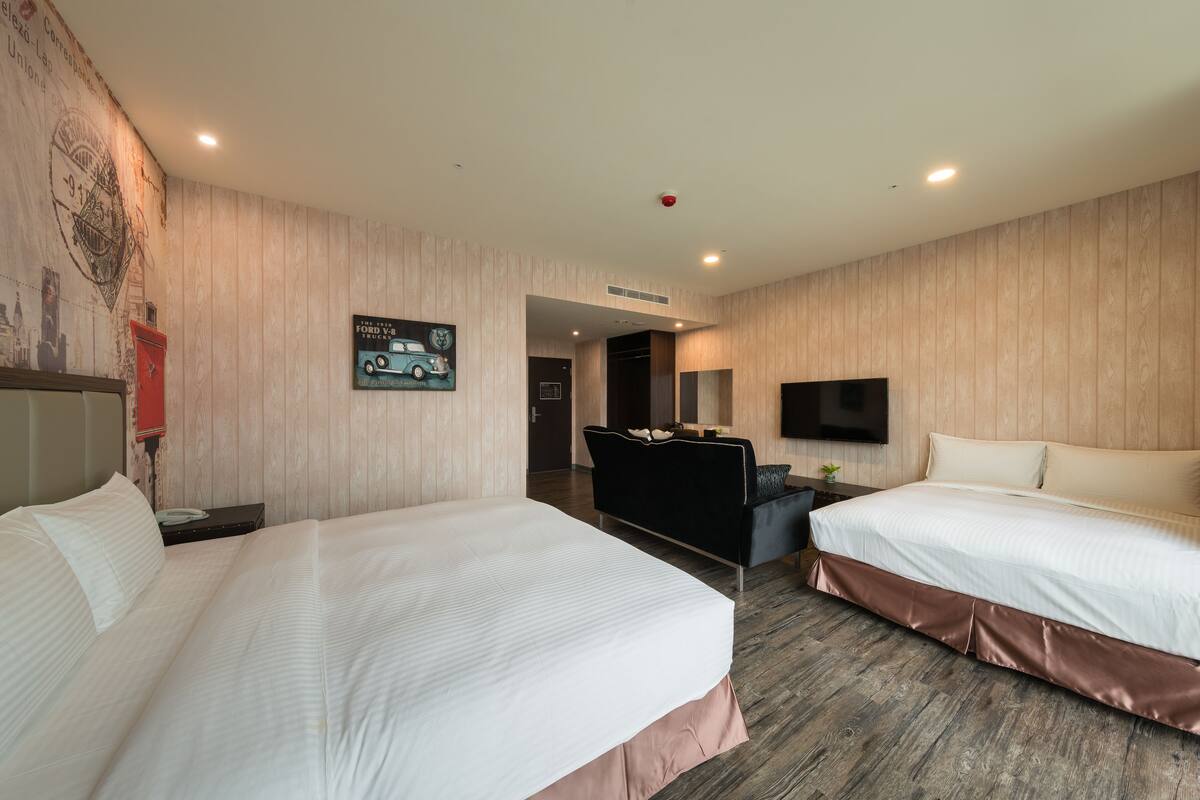 Habitación de hotel con dos camas, un escritorio, un televisor y una pared decorada con carteles publicitarios de automóviles antiguos.