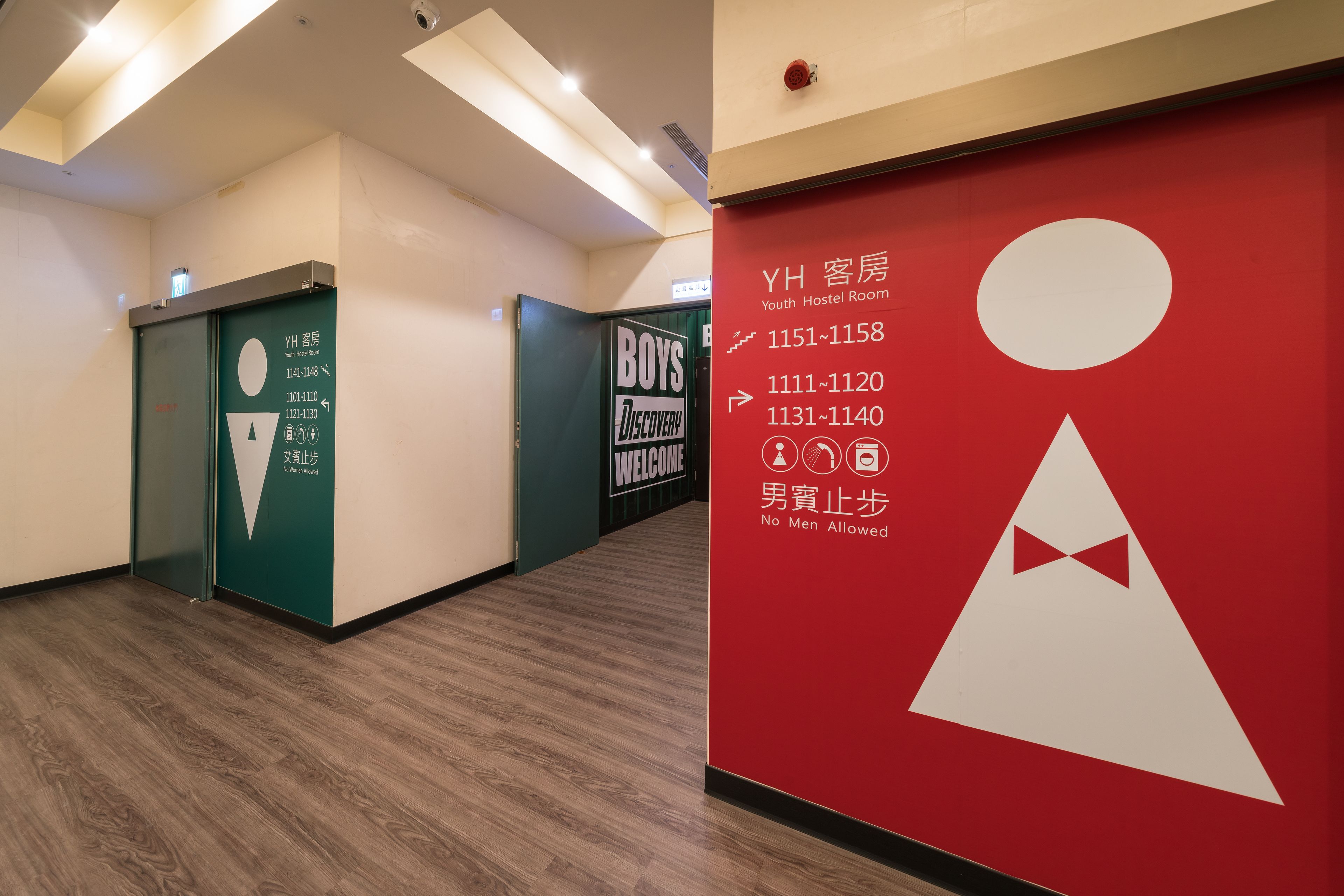 A hallway with a red wall displaying room numbers and a 'No Men Allowed' sign.