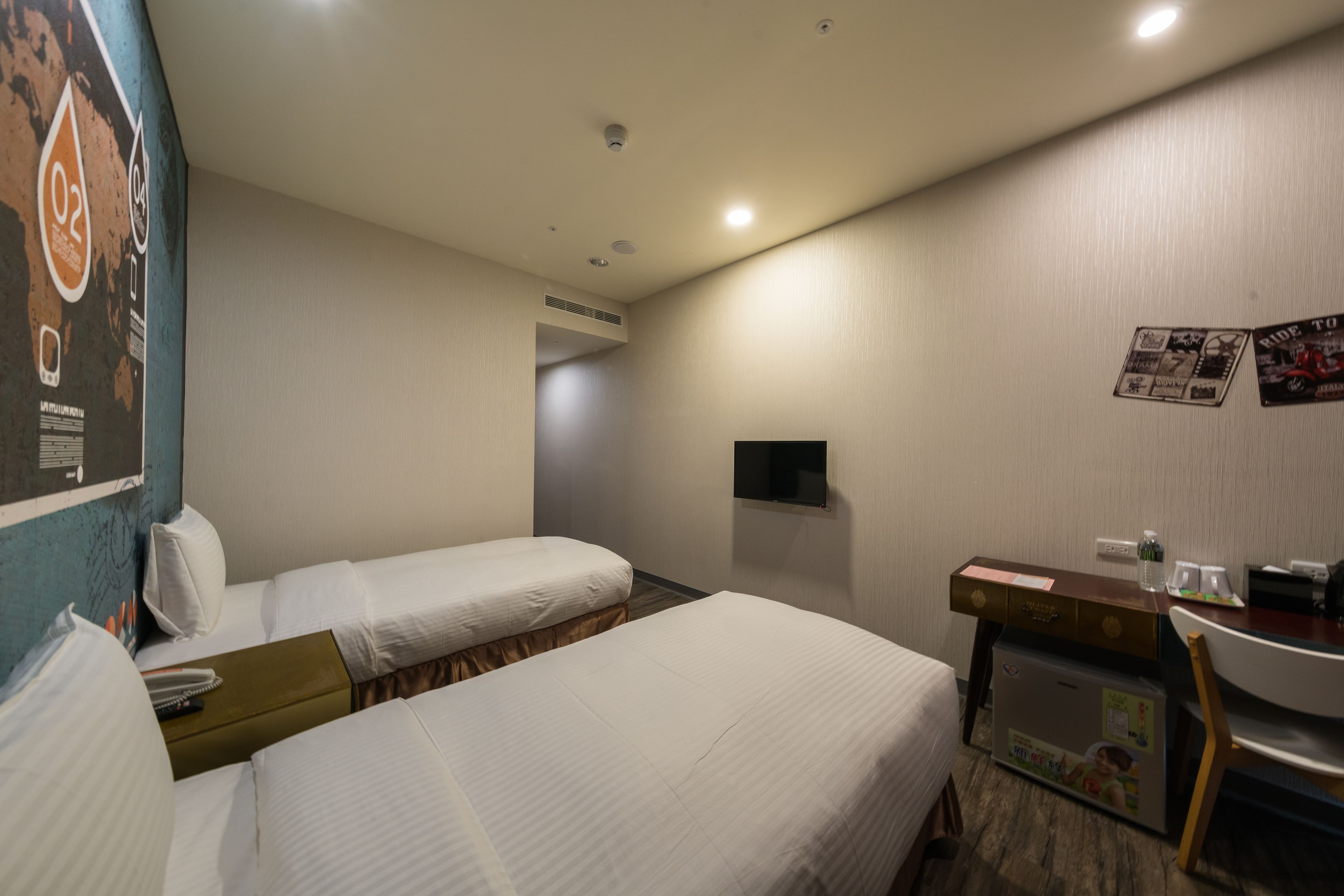 A hotel room with two beds, a desk, a chair, a TV, and a refrigerator.