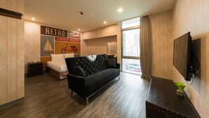 Una habitación de hotel moderna con un sofá negro, una cama con cabecero blanco, un televisor montado en la pared y un amplio ventanal con cortinas.