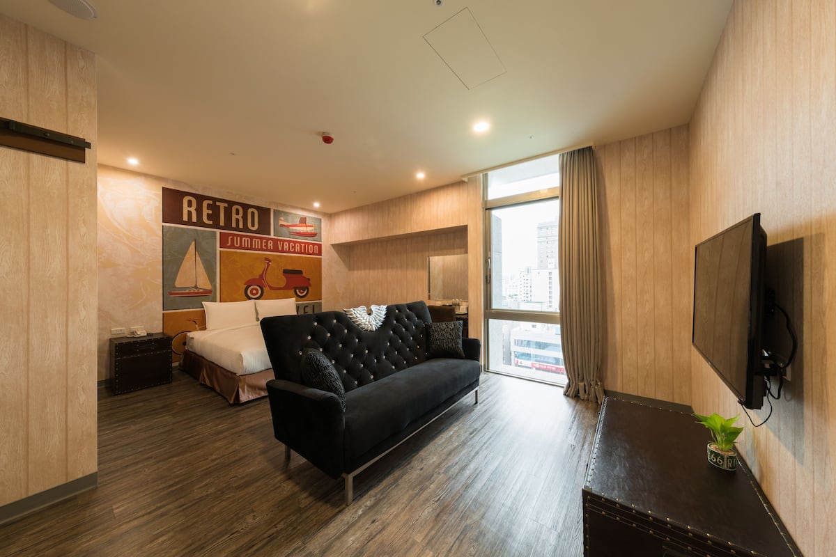 Una habitación de hotel moderna con un sofá negro, una cama con cabecero blanco, un televisor montado en la pared y un amplio ventanal con cortinas.