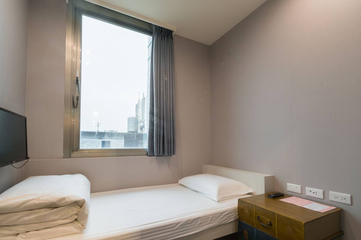 Una habitación de hotel pequeña y moderna con una cama individual, un aparador de madera, una ventana con vistas a la ciudad y un televisor montado.