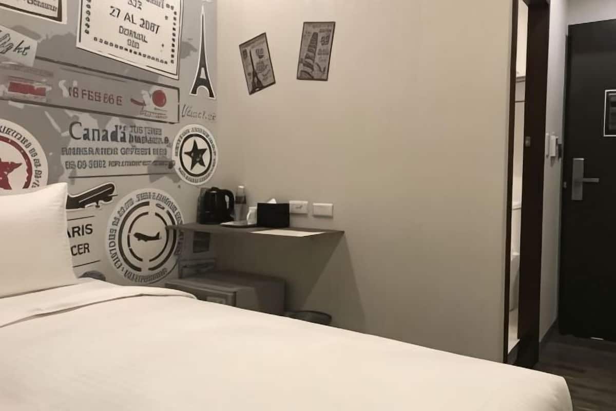 Habitación de hotel con una cama, un pequeño escritorio con silla, una mesita de noche y una pared decorada con varios adhesivos.